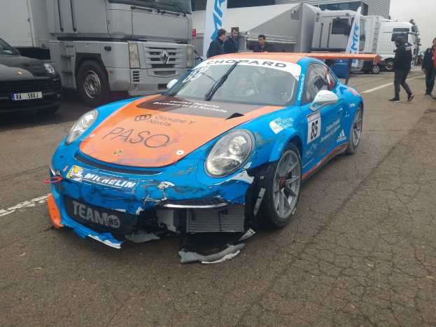 Blauer und oranger Porsche 911 GT3 RSR krachte in einen Lastwagen am Straßenrand mit Menschen in der Nähe, vor Gebäuden, Bäumen und einem klaren blauen Himmel.
