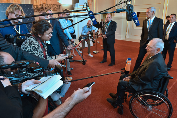 Ein Mann im Rollstuhl mit Anzug und Krawatte spricht zu Reportern mit Kameras und Mikrofonen, mit einem Banner und einer Wand im Hintergrund.
