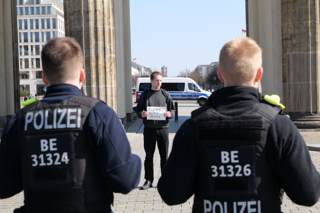 Drei Polizeibeamte stehen vor einem Gebäude, ein Mann in der Mitte hält ein Papier, flankiert von Säulen; Fahrzeuge, Menschen, Bäume und Gebäude sind im Hintergrund zu sehen.