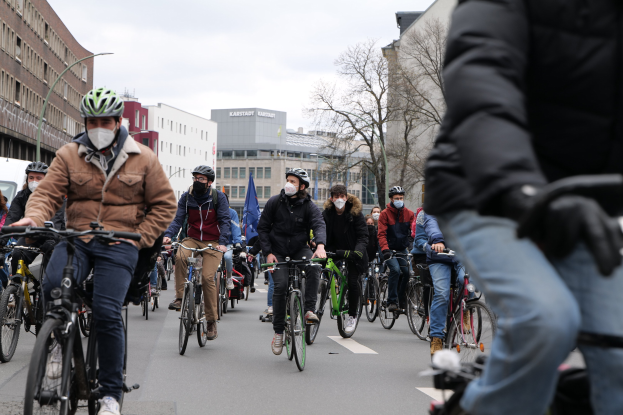 Gruppe von Radfahrern in Helmen und Handschuhen auf einer von Bäumen gesäumten Straße in Berlin fahrend, mit Gebäuden, geparktem Fahrzeug und Himmel im Hintergrund.