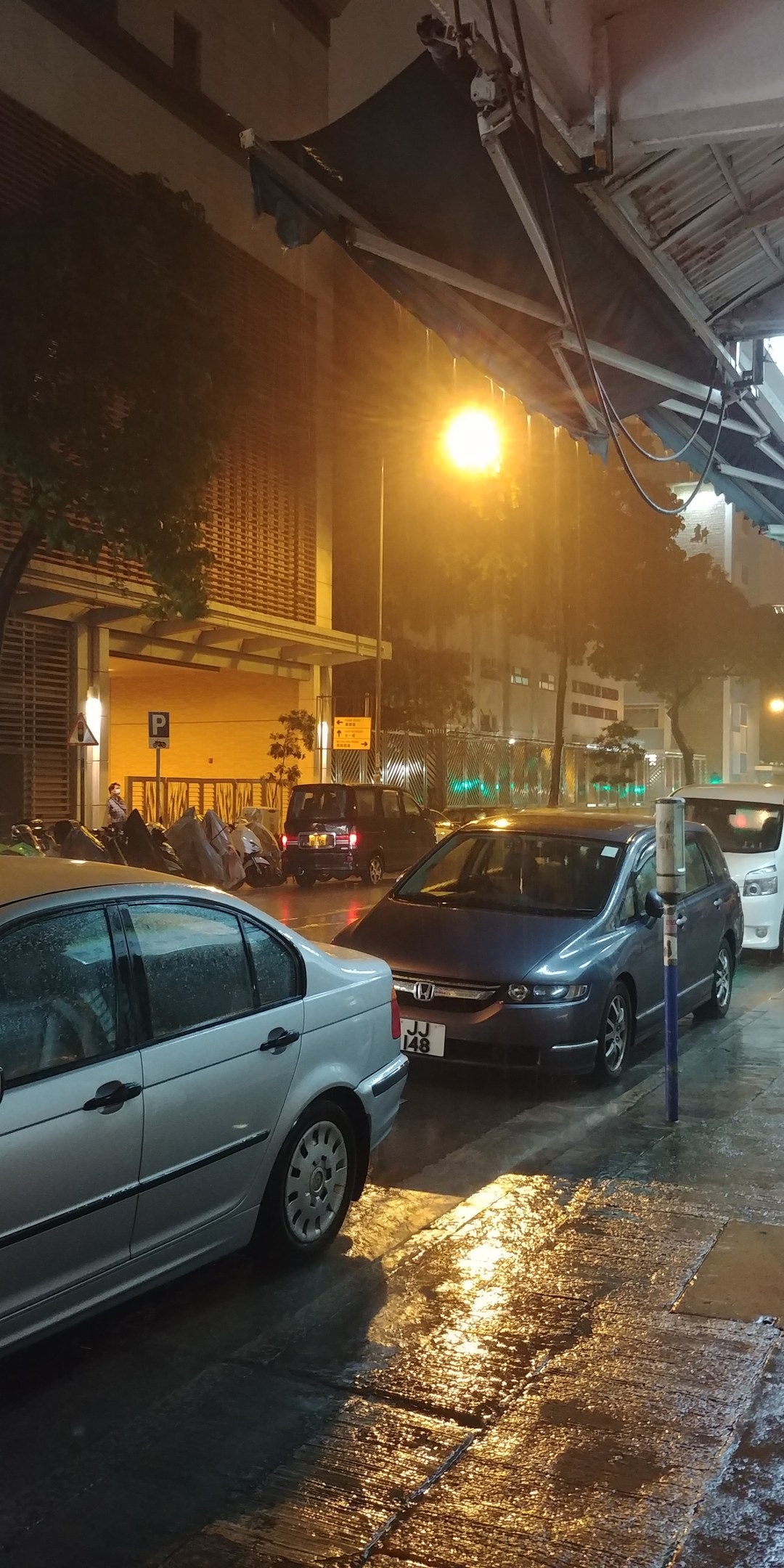 Eine nächtliche Stadtstraße mit parkenden Autos, beleuchtet von Straßenlaternen, mit einem Van, Gebäuden und Bäumen im Hintergrund.