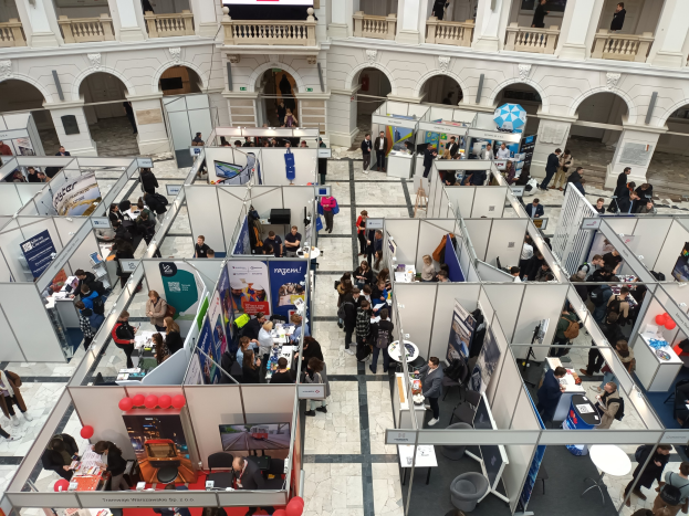 Große Besuchermenge auf einer internationalen Fachmesse mit Ständen, Bannern, Tischen, Stühlen und Ballons in einem Raum mit architectural arches, Säulen und einem Bildschirm im Hintergrund.