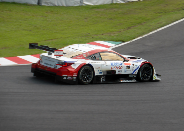Ein Sportwagen mit Text und Zahlen, der bei den 24 Stunden von Le Mans teilnimmt, fährt auf einer Rennstrecke mit Gras auf beiden Seiten und einem Zaun im Hintergrund.