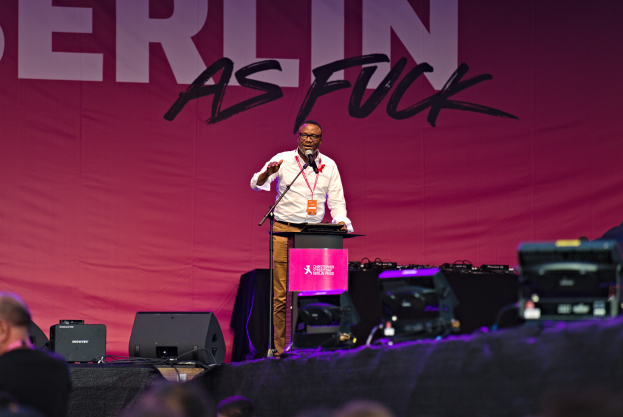 Mann in weißem Hemd und braunen Hosen hält Mikrofon auf Bühne, pinker Banner 'Berlin as Fuck' im Hintergrund