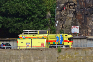 Rettungswagen auf der Seite einer Straße neben einer Klippe geparkt, mit einer Wand, einem Geländer, Pfosten, Lampen, Schildern mit Text und anderen Gegenständen im Vordergrund und Bäumen und einem Auto im Hintergrund.
