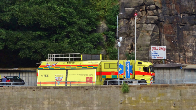 Rettungswagen auf der Seite einer Straße neben einer Klippe geparkt, mit einer Wand, einem Geländer, Pfosten, Lampen, Schildern mit Text und anderen Gegenständen im Vordergrund und Bäumen und einem Auto im Hintergrund.
