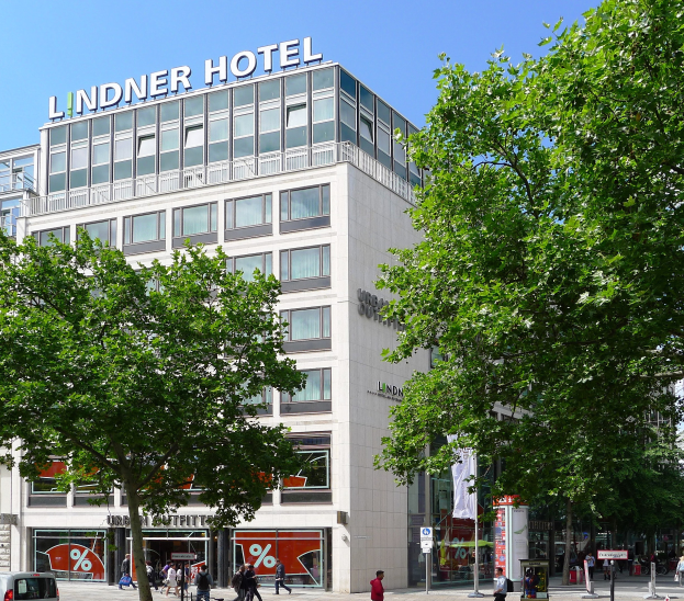 Großes modernes Hotelgebäude mit Glasfenstern, der Name "Lindner" auf dem Dach, umgeben von Bäumen und einer belebten Straße mit Fußgängern und Fahrzeugen.