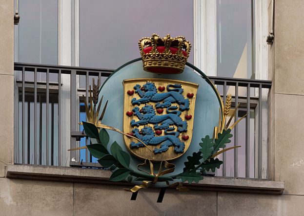 Das Wappen der britischen Königsfamilie auf der Fassade eines Gebäudes, neben einer Krone und einem Blätter-Logo, mit Fenstern und Metallgittern im Hintergrund.