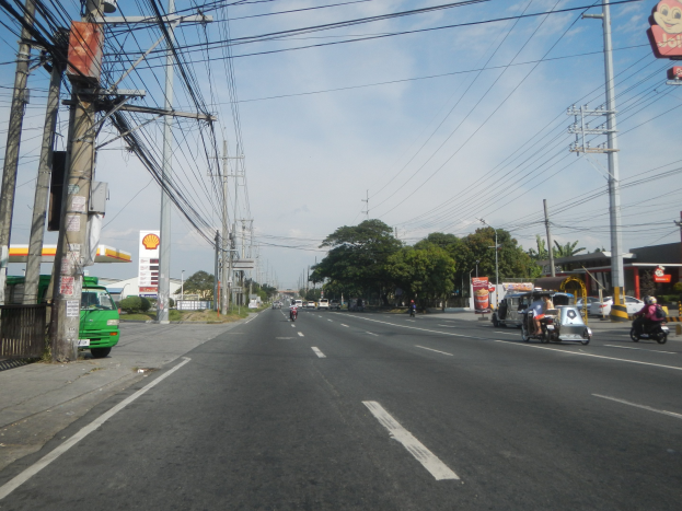 Stadtstraße mit elektrischen Polen, Drähten, Fahrzeugen, einer Tankstelle auf der linken Seite, Bäumen, Gebäuden und einem bewölkten Himmel.
