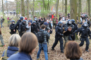 Eine Gruppe von Menschen in Einsatzkleidung steht vor einer Menge mit Helmen und Taschen, mit Bäumen, einer Fahne und Gebäuden im Hintergrund und Blättern auf dem Boden.
