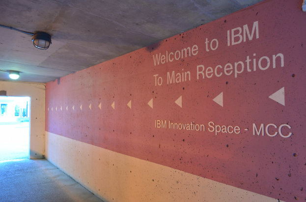 Eine Wand mit dem Text "Willkommen bei IBM", beleuchtet von Deckenlampen, mit einer Tür im Hintergrund, die "IBM Innovation Space MCC" markiert.