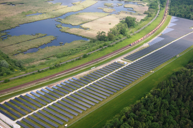 Luftaufnahme einer Solar-Farm mit Solar-Panelen auf einem Feld, umgeben von Bäumen, Gras, Pflanzen und Wasser, mit einem Zug auf einer nahen Bahnstrecke.