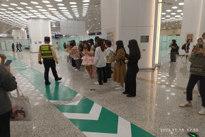Eine Gruppe von Menschen steht in einer Reihe an einem Flughafen-Check-in, einige halten Taschen und Mobiltelefone, mit Säulen im Hintergrund und Lampen an der Decke und einem Wasserzeichen am unteren Rand des Bildes.