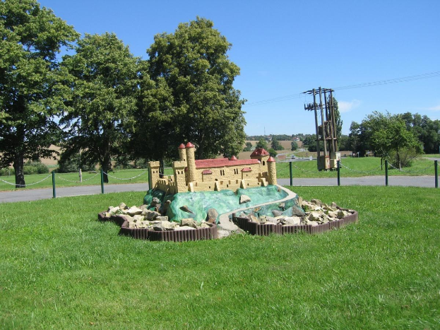 Minigolfplatz mit einem grasbewachsenen Feld, umgeben von einem Zaun mit Pfählen und Ketten, Bäumen, Strommasten mit Drähten und einem bewölkten Himmel im Hintergrund.