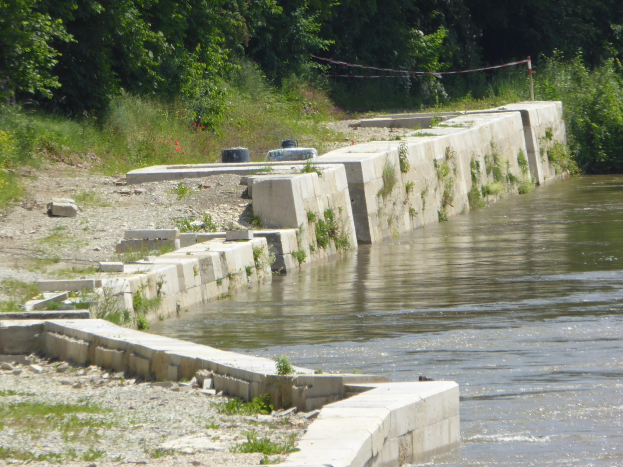 Ein ruhiger, klarer Fluss mit einer zentralen Rückhaltewand, umgeben von üppiger Vegetation und Bäumen, die den Himmel und das Laub spiegeln.
