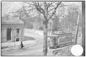 Ein Schwarz-Weiß-Foto einer Straßenbahn auf einer Straße in der Stadt, mit einer Person, die ein Fahrrad vor ihr fährt, einigen Leuten in der Straßenbahn und Bäumen und einem Gebäude im Hintergrund.