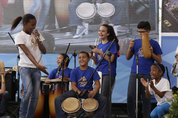Kinder auf der Bühne mit einer Frau mit Mikrofon, umgeben von Musikinstrumenten und Mikrofonstativen, mit einem Banner und Pflanzen im Hintergrund.