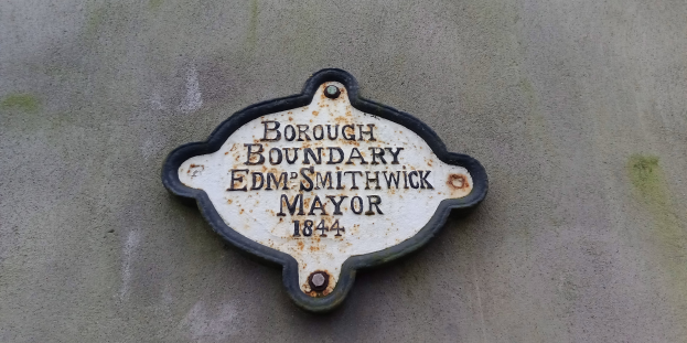 Metallplatte an einer Gebäudeseite mit schwarzem Hintergrund und weißer fettgedruckter Schrift, die "Stadtteilgrenze Edm Smithwick Bürgermeister" lautet.