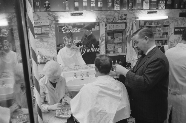 Schwarz-weiß-Foto eines Mannes, der einer Frau die Haare schneidet in einem Friseursalon mit Waschbecken, Spiegel und Regalen im Hintergrund.