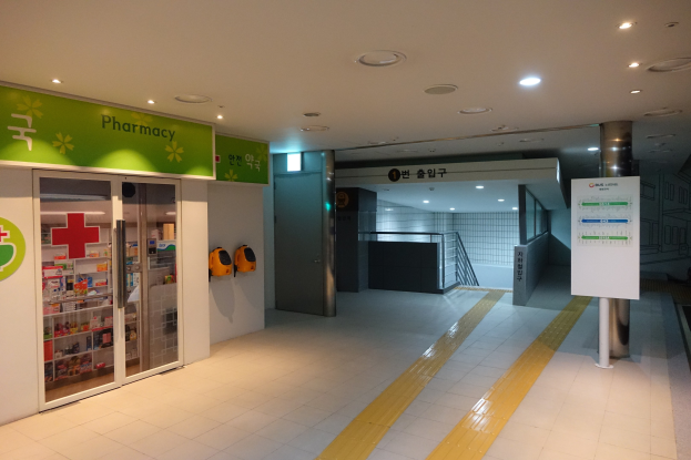 Apothekeneingang mit einem "Apotheke"-Schild auf der linken Seite, einer Tafel mit Text auf der rechten Seite, Hintergrundobjekten und beleuchteter Dachbeleuchtung.