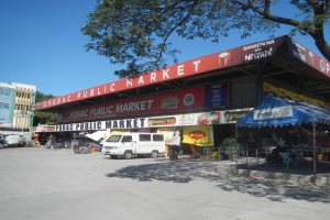 Öffentlicher Markt mit einem vor ihm geparkten Van, umgeben von Gebäuden, Geschäften, Schildern, Fahrzeugen auf der Straße, Menschen unter einem Zelt sitzend, Geländern, Bäumen und einem klaren blauen Himmel.