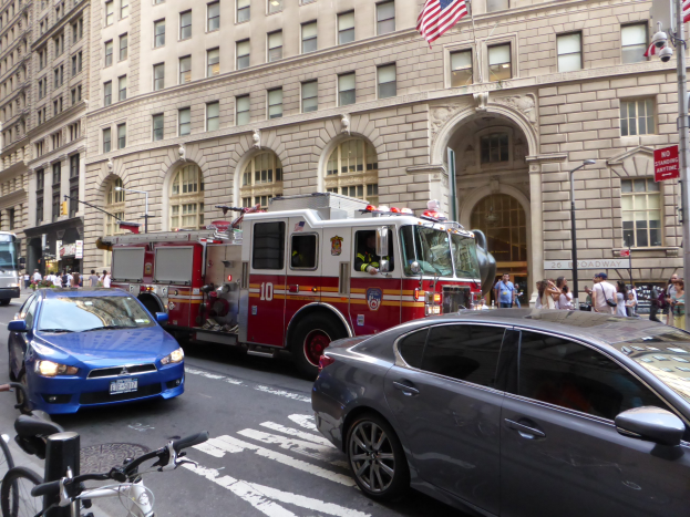 Ein rotes Feuerwehrauto auf einer Stra├če in New York City mit fahrenden Fahrzeugen, Passanten, parkenden Fahrr├Ądern und Geb├Ąuden im Hintergrund.