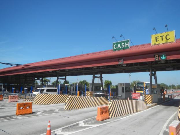 Mautstation mit einem "Bar"-Schild, Fahrzeugen, Absperrpfeilern, Schildern, Straßenlaternen, Kabeln, Bäumen und einer bewölkten Himmel.