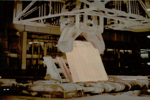 Altes Foto eines großen Industriekrans, der ein Metallförderband in einer Fabrik hochhebt, mit einem Gebäude im Hintergrund.