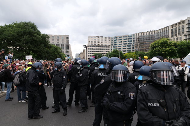 Große Gruppe von Polizeibeamten vor einer Kamera schwingenden Menge auf einer Baumgesäumten Straße in Berlin, mit Gebäuden und einem bewölkten Himmel im Hintergrund.