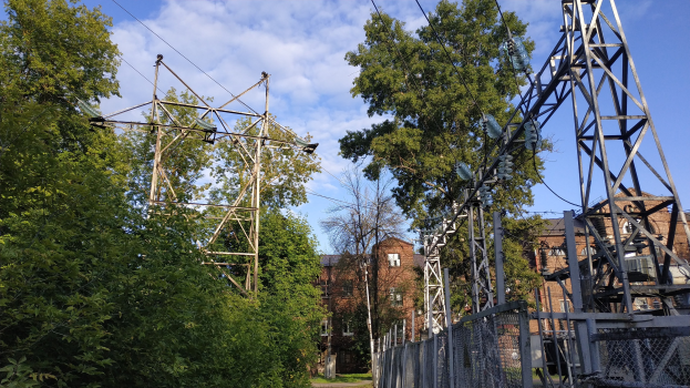 Gro├če Strommast mit mehreren Dr├Ąhten, die durch einen bewaldeten Bereich verlaufen, umgeben von B├Ąumen, Masten, Z├Ąunen und einem Zaun, mit im Hintergrund sichtbaren Geb├Ąuden unter einem bew├Âlktem Himmel.