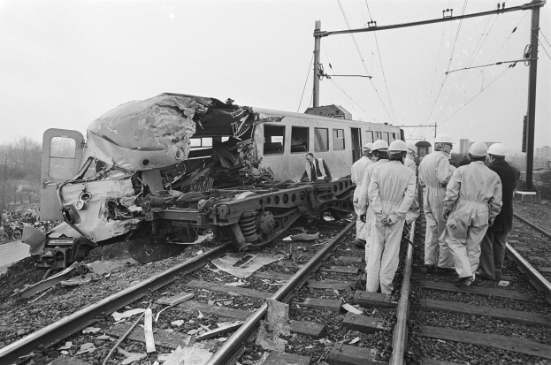 Ein Schwarz-Wei"s-Bild einer Zugkollision auf Gleisen mit Menschen in Helmen in der N"he, umgeben von Strommasten, B"umen, Geb"uden und einem klaren Himmel.