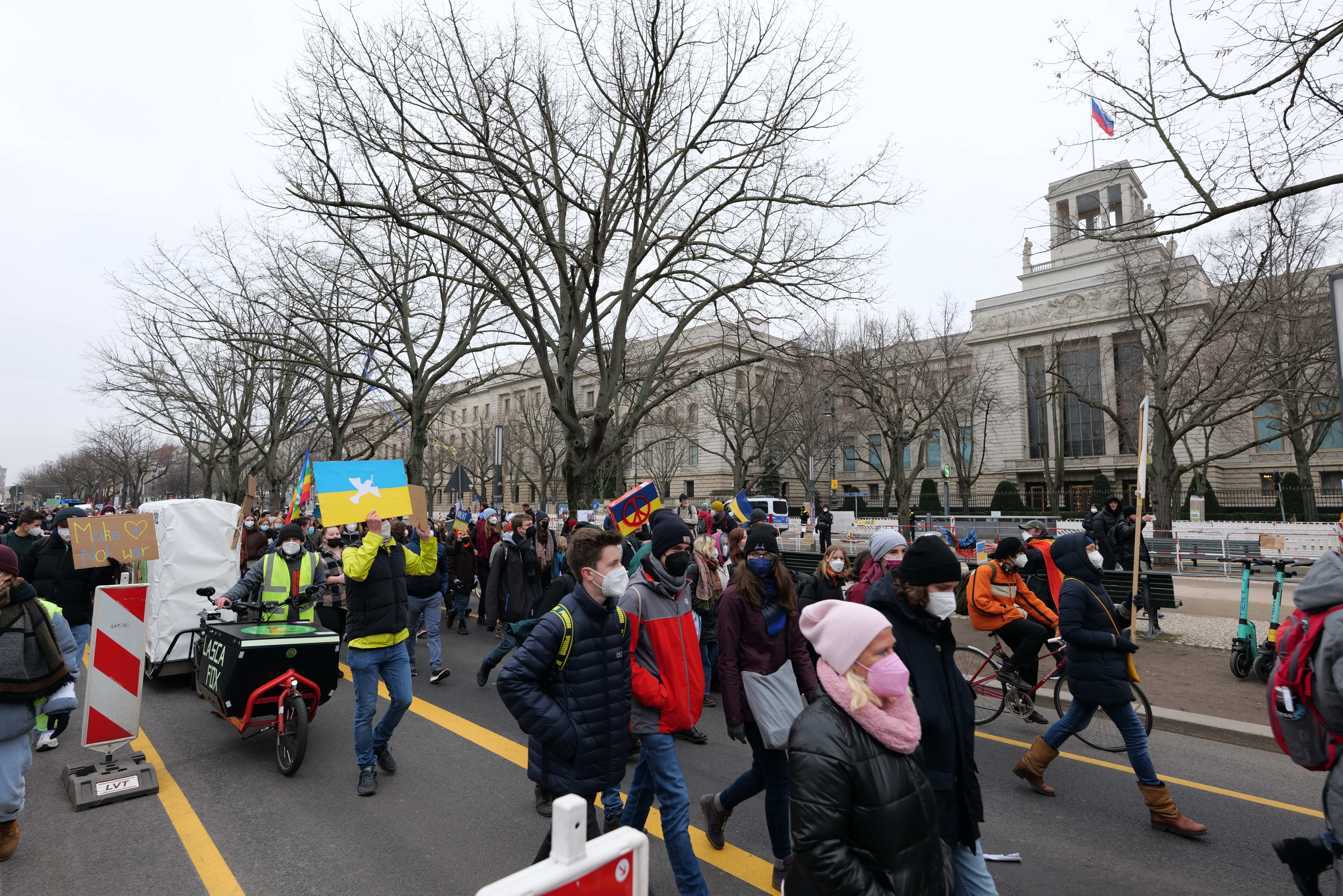 Eine große Gruppe von Menschen nimmt an einer Protestdemo auf einer Straße in Washington, D.C. teil, einige halten Schilder und Transparente, andere fahren Fahrräder, und es gibt Schilder, Bäume und einen klaren blauen Himmel im Hintergrund.