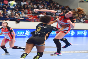 Eine Gruppe von Frauen spielt Handball auf einem Court mit einem Ball in der Luft, während Zuschauer von Stühlen, Geländern und einer Wand im Hintergrund zusehen.
