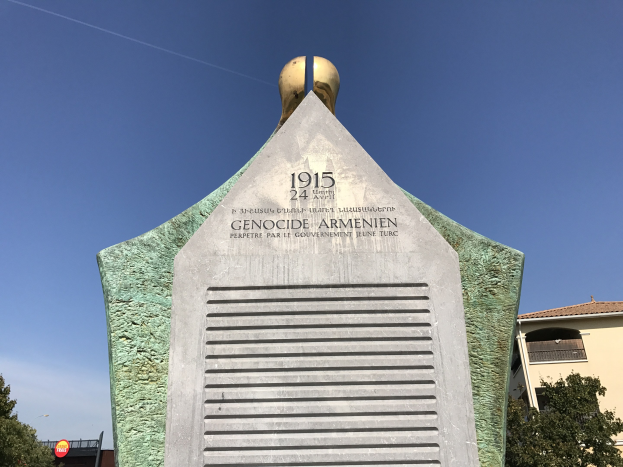 Ein hoher Denkmal mit einer Uhr darauf, mit Text beschriftet, steht in einem Park umgeben von Bäumen und Gebäuden unter einem klaren Himmel, erinnert an den 24. Jahrestag des Völkermords in Armenien.