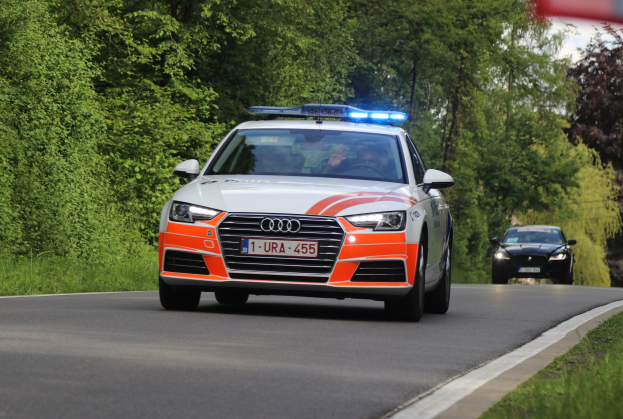 Polizeifahrzeug (Audi A4) auf grasbewachsener Straße neben einem Wald mit zwei Insassen unter einem klaren blauen Himmel.