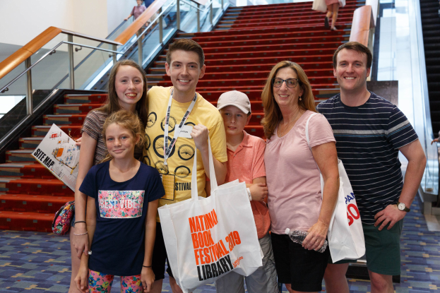 Gruppe von Menschen, die vor einer Rolltreppe an der Nationalen Buchmesse 2019 lächeln und Taschen halten, mit sichtbaren Stufen und Geländern im Hintergrund.