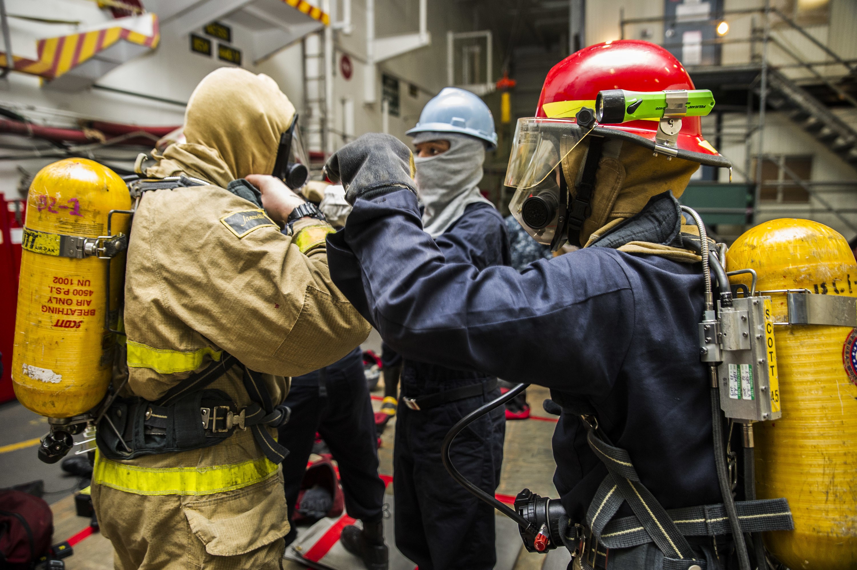 Feuerwehrleute in Schutzausrüstung mit Helmen, Handschuhen und Sauerstoffflaschen stehen zusammen, mit Equipment-Taschen auf dem Boden, neben Treppen und Geländern in einem beleuchteten Innenbereich.