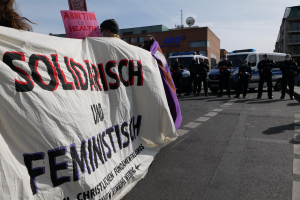 Gruppe von Menschen marschiert auf einer Straße mit einem "Solidarität und Feminismus"-Schild, mit parkenden Fahrzeugen, Gebäuden, einer Schüsselantenne und einem klaren blauen Himmel im Hintergrund.