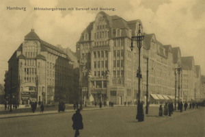 Schwarzes und weißes Foto von Menschen, die eine Straße in Hamburg, Deutschland, entlanggehen, mit Gebäuden, Straßenlaternen, Lichtern und Kraftfahrzeugen, die entlang der Straße aufgereiht sind, unter einem bewölkten Himmel.