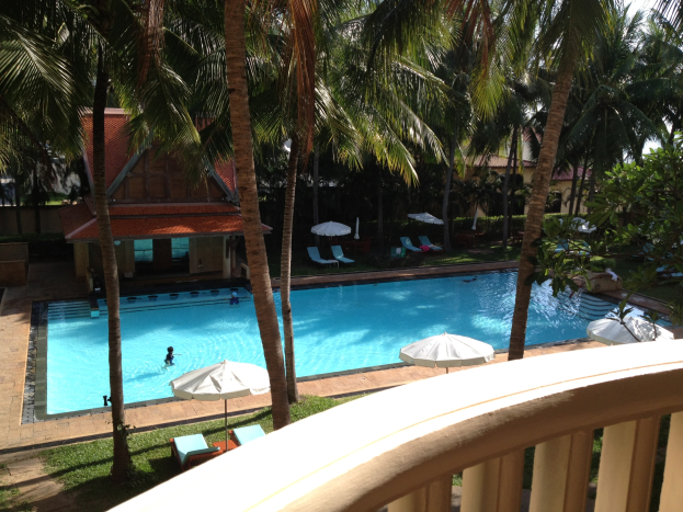 Ein großes Schwimmbad umgeben von Grün, mit Menschen, die schwimmen, Schirmen, Stühlen, Häusern im Hintergrund und einem klaren blauen Himmel, von einem Hotelbalkon aus betrachtet.