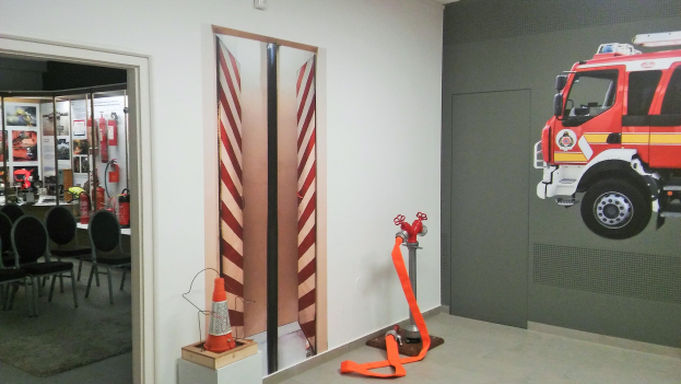 Ein Raum mit einer Wandmalerei eines Feuerwehrautos, einer Tür, einem Verkehrskegel, einem Ständer, einem Teppichboden, Stühlen, Tischen und Feuerlöschern.