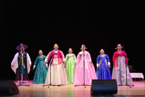 Eine Gruppe von Menschen auf einer Bühne in bunten Kleidern, die in Mikrofone mit Ständern singen, mit Lautsprechern unten und einem Tisch mit einer Decke rechts, vor einem dunklen Hintergrund.