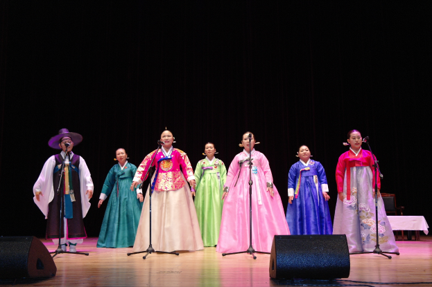 Eine Gruppe von Menschen auf einer Bühne in bunten Kleidern, die in Mikrofone mit Ständern singen, mit Lautsprechern unten und einem Tisch mit einer Decke rechts, vor einem dunklen Hintergrund.