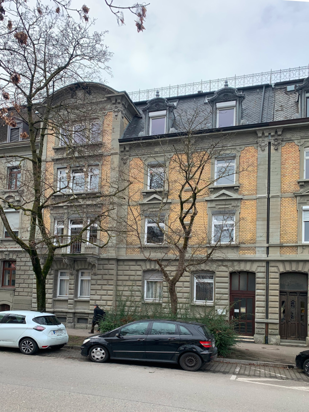 Straßenszene mit parkenden Autos, einem Fußgänger auf dem Gehweg, Grünfläche, einem mehrstöckigen Gebäude mit Fenstern und Türen und einem bewölkten Himmel, das eine Pariser Wohnung zum Verkauf zeigt.