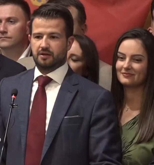 Ein Mann in Anzug und Krawatte spricht an einem Podium mit Mikrofon vor einer Menge hinter einem Banner, wahrscheinlich über ein Regierungssozialmedienverbot diskutierend.