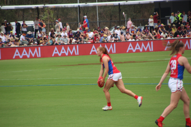 Zwei Frauen spielen Australian Rules Football auf einem Feld, eine hält den Ball, mit Zuschauern und Schildern im Hintergrund.