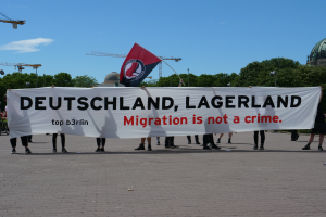 Eine Gruppe von Menschen hält ein Banner mit der Aufschrift "Deutschland, Lagerland Migration ist kein Verbrechen" und eine Flagge, steht auf dem Boden mit Straßenlaternen, Straßenlaternen, Bäumen, Kränen, einem Gebäude und einem bewölkten Himmel im Hintergrund.
