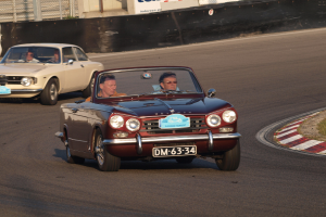 Zwei Autos auf einer Rennstrecke mit Zuschauern, umgeben von Gras und einem eingezäunten Hintergrund mit Texttafeln.