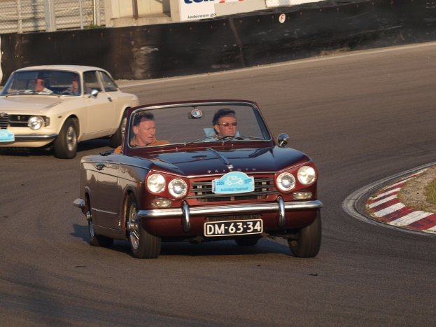 Zwei Autos auf einer Rennstrecke mit Zuschauern, umgeben von Gras und einem eingezäunten Hintergrund mit Texttafeln.