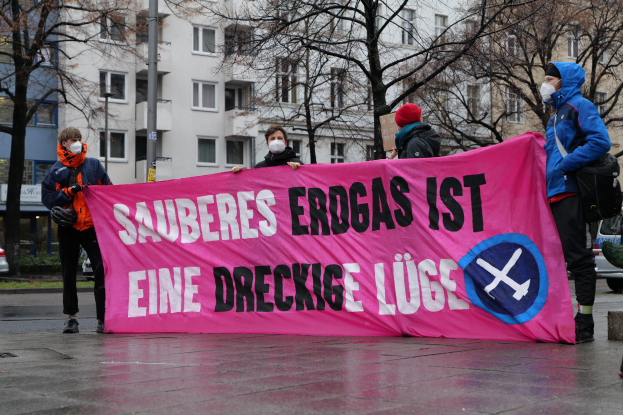 Eine Gruppe von Menschen mit einer pinken Fahne, auf der "Sauberes Erdgas ist eine Dreckige Lüge" steht, im Protest, mit Fahrzeugen, einem Schild, Bäumen, einem Gebäude und einem bewölkten Himmel im Hintergrund.